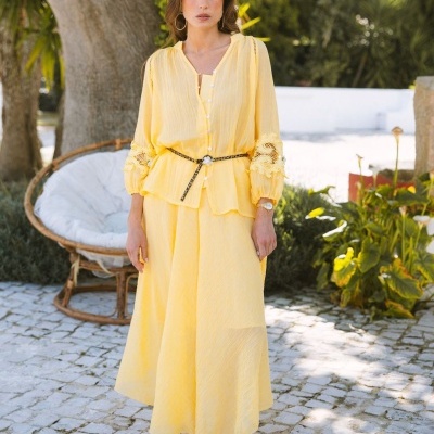 Conjunto feminino amarelo com blusa de botões e saia longa, sandálias castanhas, ao ar livre em ambiente rústico