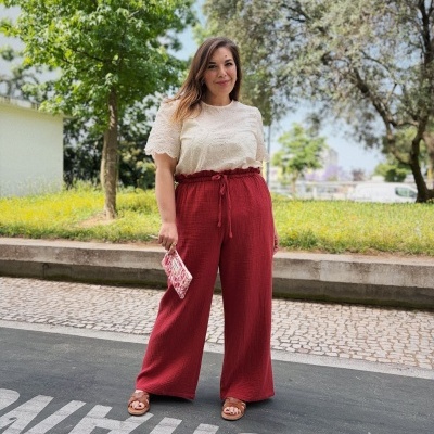 Mulher com blusa branca bordada, calças largas bordô, sandálias castanhas e carteira pequena com padrão floral.