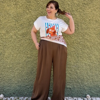 Pessoa com T-shirt branca e calças castanhas largas contra parede verde texturada.