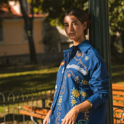 Camisa azul bordada com padrões florais e geométricos coloridos, com renda branca, usada ao ar livre