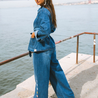 Mulher com casaco e calças de denim azul e bordados florais ao lado de um lago, com ponte ao fundo.