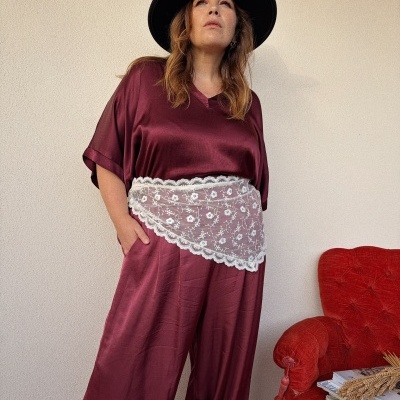 Mulher com vestido vinho acetinado, lenço branco em renda na cintura e chapéu preto junto a uma poltrona vermelha