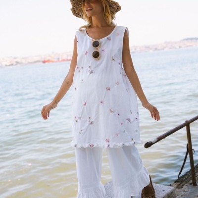 Vestido branco com bordados coloridos, calças largas brancas, chapéu de palha castanho e sandálias castanhas junto ao mar.