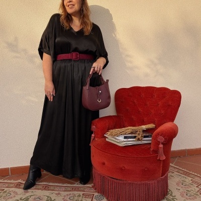 Mulher com vestido preto, cinto vermelho, chapéu preto e mala vinhos ao lado de poltrona vermelha