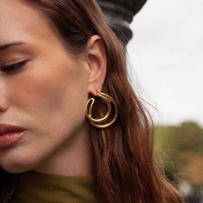 Brincos de argolas douradas e colar dourado em mulher com top verde-oliva ao ar livre