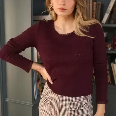 Mulher com camisola vinho e calças xadrez pois está à frente de uma estante de livros.