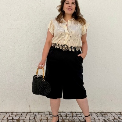 Mulher com camisa creme rendada e calças pretas com cinto largo, sandálias pretas e mala preta com alças de bambu.