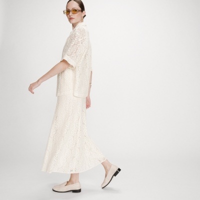 Mulher vestindo conjunto branco de renda com saia comprida e camisa de mangas curtas, usando óculos com lentes amarelas e sapatos bege.