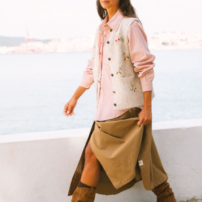 Moda feminina com colete bordado, camisa rosa, saia caqui e botas castanhas ao ar livre junto à água