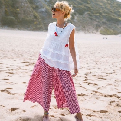 Mulher com blusa branca e calças xadrez vermelhas na praia com acessórios coloridos e óculos de sol.