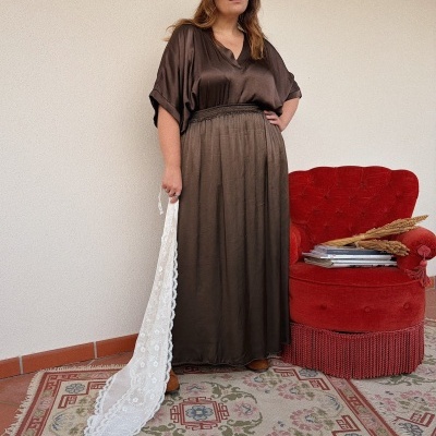 Mulher com vestido castanho e chapéu segurando tecido rendado em ambiente com poltrona vermelha e tapete estampado