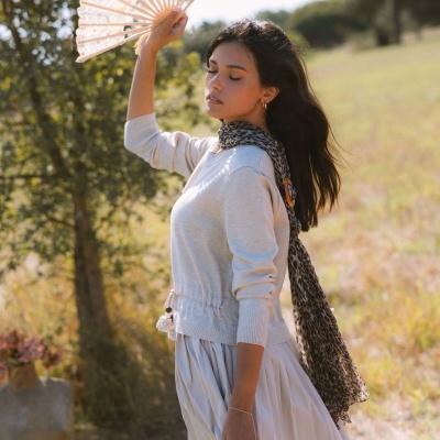 Mulher com blusa creme e saia branca plissada, lenço animal print e leque branco ao ar livre