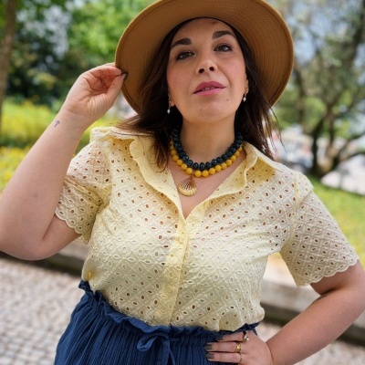 Mulher com blusa amarela, calças ou saia azul, chapéu castanho, colar e joias