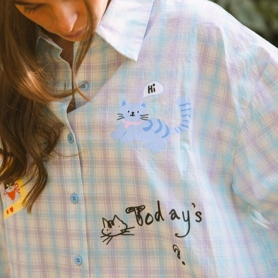 Camisa xadrez pastel com estampas de gatos, flores e texto, vista de perto com luz natural