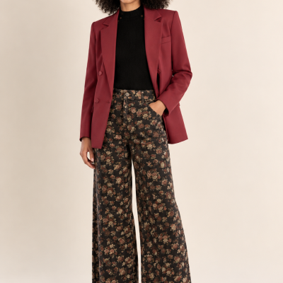 Mulher com blazer bordô, camisola preta, calças florais largas e sapatos pretos em fundo neutro