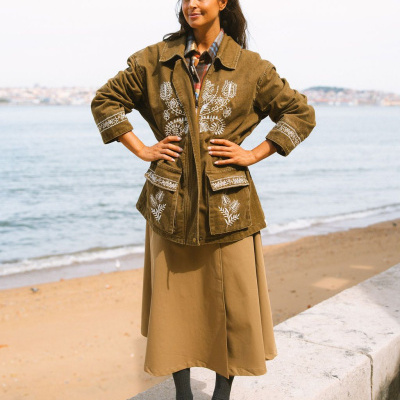 Mulher com casaco castanho bordado, saia comprida, camisa às riscas, meias e sapatos castanhos junto ao mar