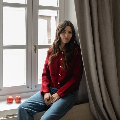 Mulher sentada junto a uma janela com blusa vermelha de botões dourados, calças de ganga azuis e cortina bege ao lado.