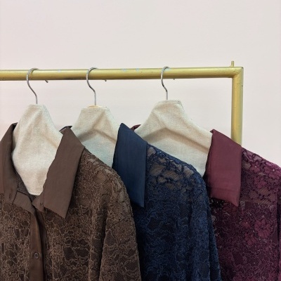 Três camisas de renda em castanho, azul escuro e bordô penduradas em cabides numa estrutura metálica dourada