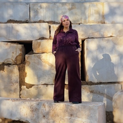 Mulher com calças castanhas largas e blusa de padrão floral violeta, usando bandana rosa, em cenário de blocos de pedra