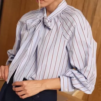 Blusa azul claro listrada com laço na gola e calças escuras, em ambiente de escritório