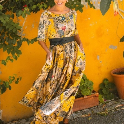 Vestido comprido amarelo com padrão floral e cinto preto junto a parede amarela com plantas