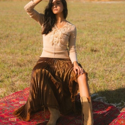 Mulher com suéter bege, saia bronze plissada e botas bege sentada num tapete vermelho num campo