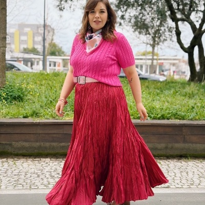 Mulher com camisola rosa, saia vermelha plissada e sandálias em rua urbana