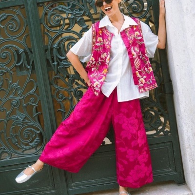 Mulher com colete rosa bordado, camisa branca e calças largas rosa com padrão floral, com óculos escuros e sapatos prateados em porta verde ornamentada.