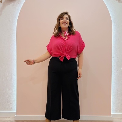 Mulher com camisa rosa e calças pretas em arco rosa claro com parede branca.