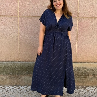 Vestido azul-marinho com bolinhas brancas e sandálias pretas, mulher sorrindo com parede cor de rosa ao fundo