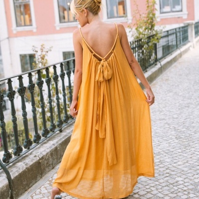 Vestido longo amarelo mostarda com alças finas e laço nas costas, usado por uma mulher de costas numa rua com calçada de pedra