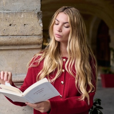 Mulher a ler livro com casaco vermelho e calças de ganga junto a parede de pedra