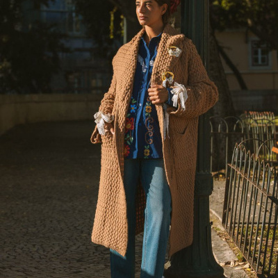 Mulher com casaco castanho de malha, blusa azul com bordados florais e calças de ganga em rua com calçada portuguesa