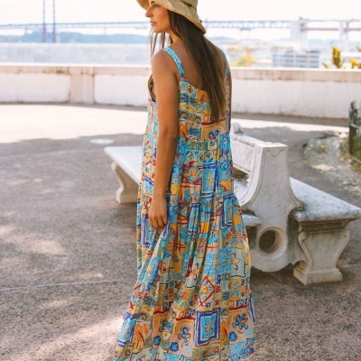 Mulher de costas com vestido colorido longo e chapéu de palha, em área exterior com banco de pedra e ponte ao fundo.