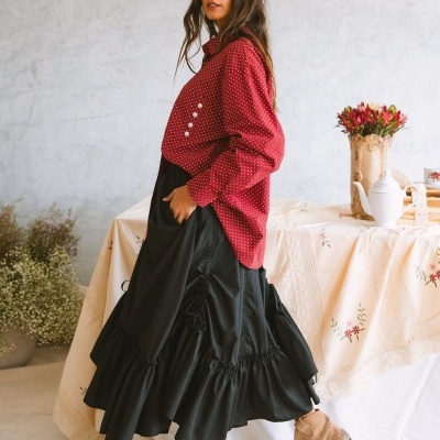Mulher com camisa vermelha de bolinhas brancas, saia preta folhada e botas castanhas em ambiente interior decorado.