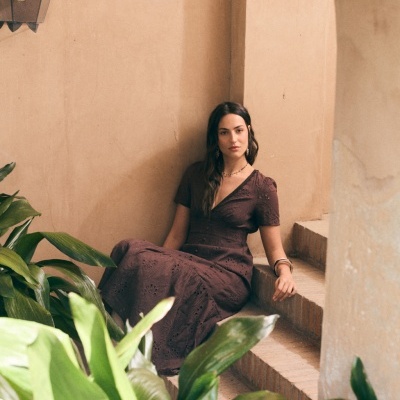 Vestido castanho com padrão floral recortado, usado por mulher sentada em escadas com parede bege e plantas verdes à frente