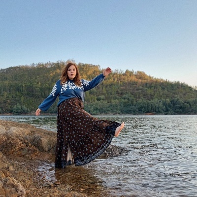 Mulher com vestido longo castanho floral e suéter azul com padrão branco junto a água e pedras com colinas ao fundo