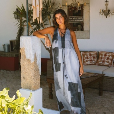 Mulher com vestido comprido cinzento e branco, sandálias de cunha, num pátio com pilar de pedra e móbile de vento de madeira.