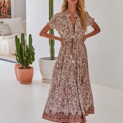 Vestido comprido feminino com padrão floral em tons rosa e vermelho, com folhos e laço na cintura, usado por mulher no interior com plantas