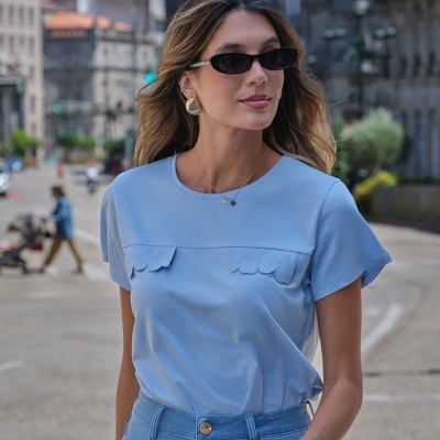 Mulher a usar t-shirt azul clara, jeans azul, óculos de sol pretos e joias douradas numa rua urbana