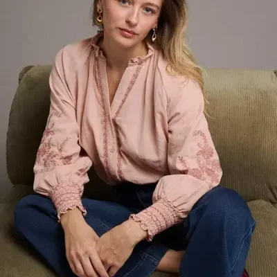 Mulher sentada num sofá verde-claro a usar blusa rosa clara bordada e jeans azul escuro