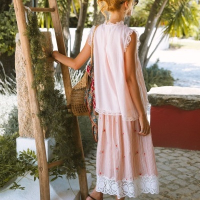Mulher com roupa clara de verão, saia e top rendados, sandálias castanhas, mochila de palha, junto a escada de madeira decorada com vegetação.