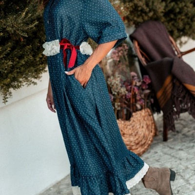 Vestido comprido azul com pontos brancos, cinto vermelho e botas castanhas em cenário exterior.
