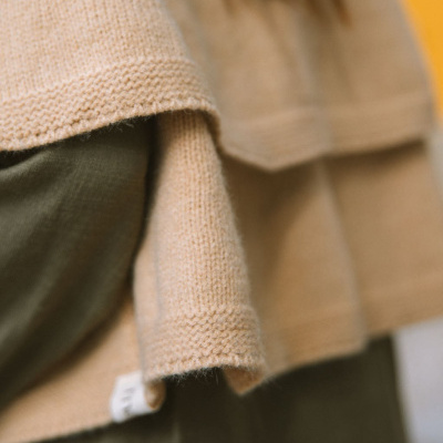 Casaco de malha bege sobre roupa verde-oliva com etiqueta branca e fundo amarelo
