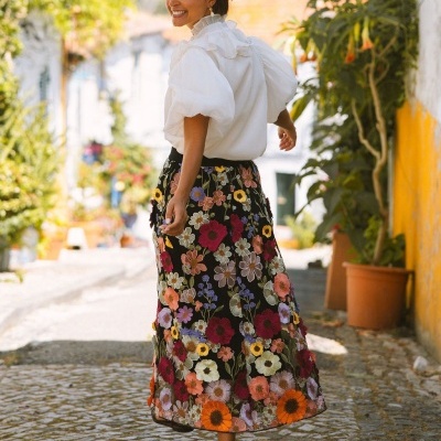 Saia preta bordada com flores coloridas usada por mulher com blusa branca de mangas bufantes.