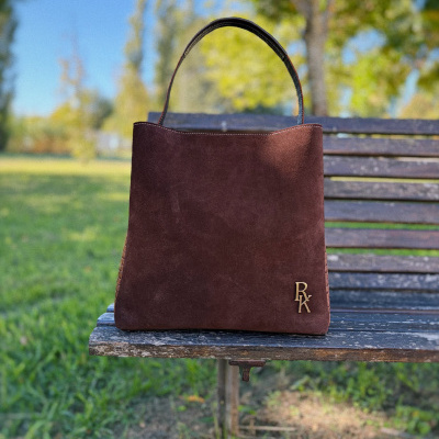 Mala de mão castanha em camurça com alça e detalhe metálico dourado RX no banco de madeira