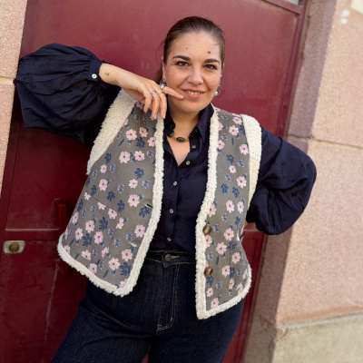 Mulher com colete floral cinzento com detalhes felpudos e camisa preta em frente a porta vermelha
