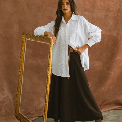 Mulher com camisa branca bordada e saia castanha longa, segurando moldura dourada