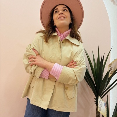 Mulher com casaco bege, camisa rosa às riscas, chapéu cor-de-rosa e calças de ganga, em fundo rosa com planta verde