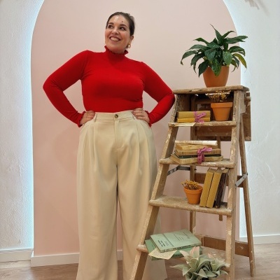 Mulher com camisola vermelha e calças bege junto a escada de madeira decorativa com plantas e livros
